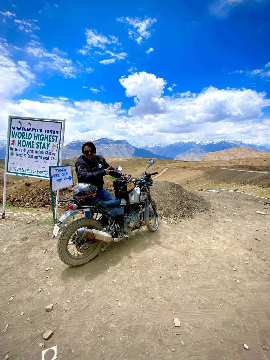 A rider on a Royal Enfield beside a sign for the world's highest homestay, blue sky and bare mountains behind