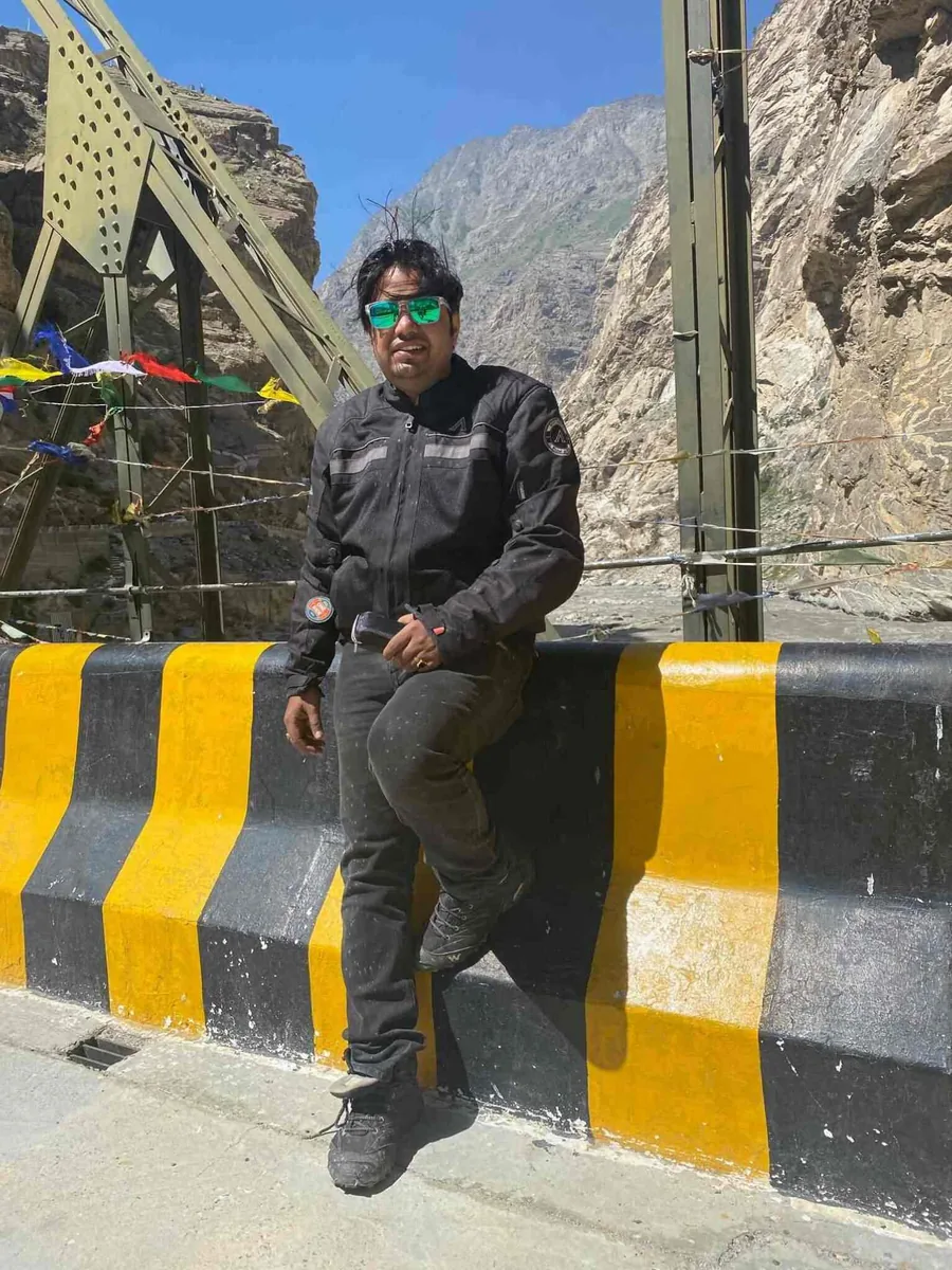 The owner in riding gear sitting on a yellow-and-black road barrier below a rock face with prayer flags
